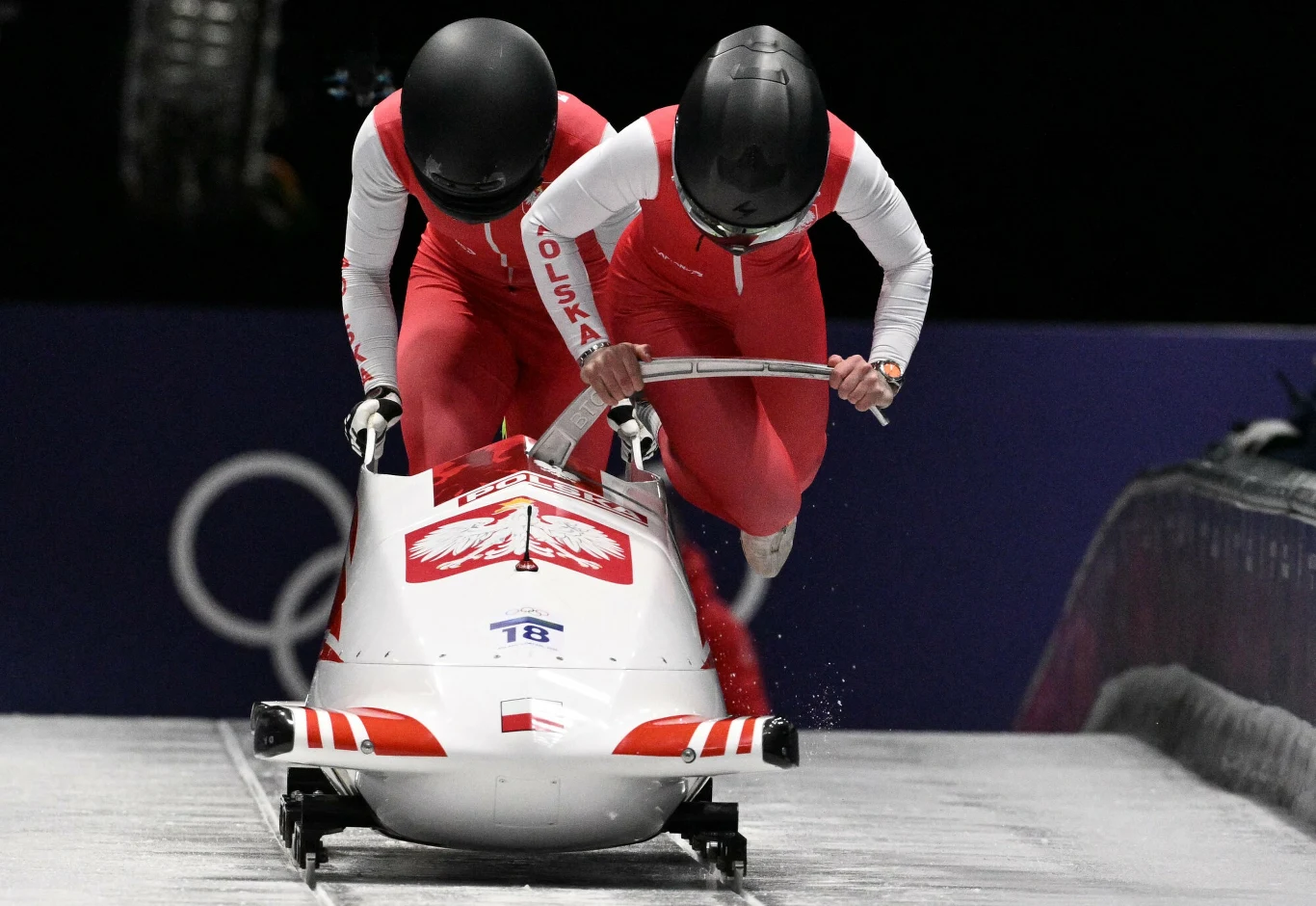 Linda Weiszewski, Klaudia Adamek Dwie osoby w czerwono-białych kombinezonach i kaskach startują do wyścigu bobslejowego na torze lodowym, prowadząc biały bobslej z polskim godłem i napisem 'Polska' na rękawach zawodników.