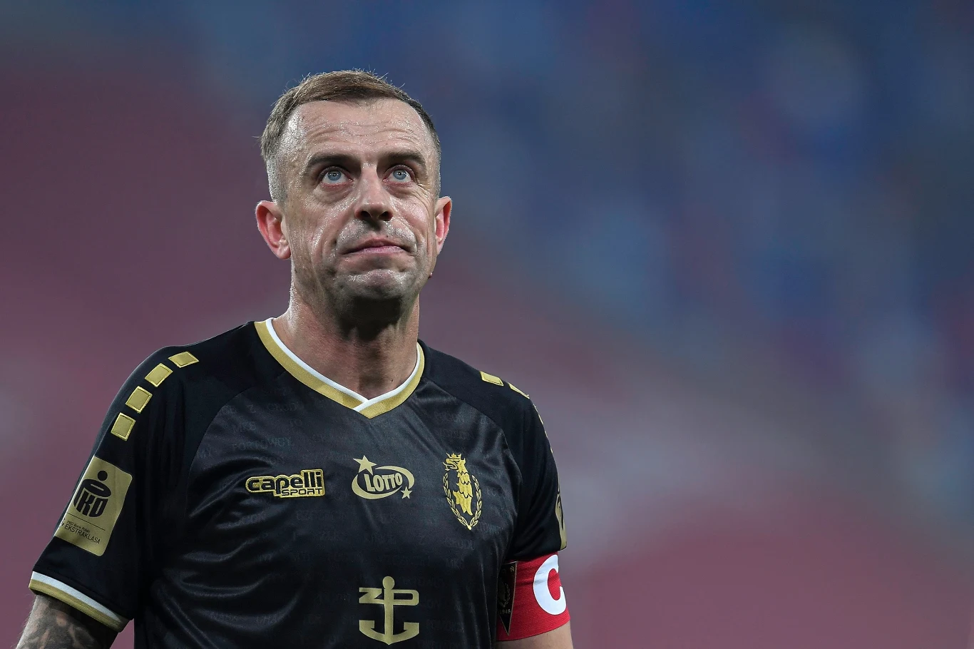 Kamil Grosicki Zbliżenie na piłkarza w czarnej koszulce z żółtymi detalami i opaską kapitana, skupionego podczas meczu.
