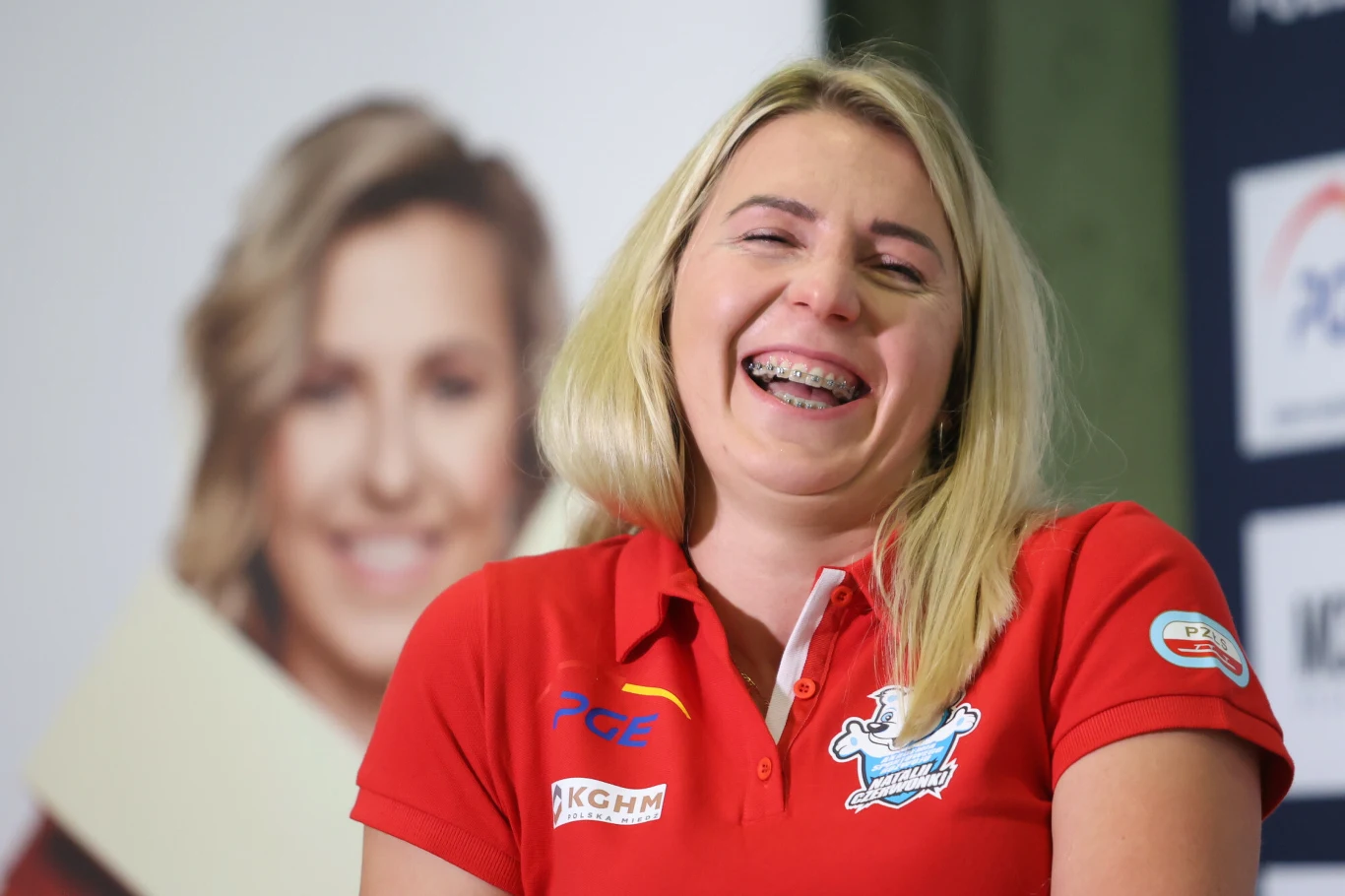 Uśmiechnięta kobieta w czerwonej koszulce sportowej z naszywkami sponsorów, w tle rozmazany plakat z portretem innej kobiety.