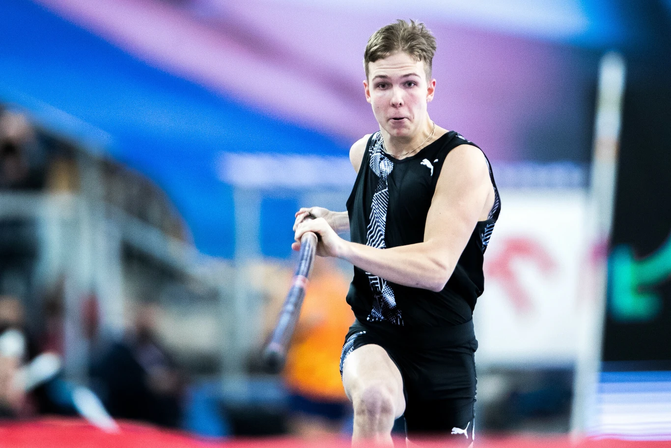 Młody mężczyzna w sportowym stroju podczas biegu z tyczką w hali lekkoatletycznej, skupiony wyraz twarzy, dynamiczne ujęcie w trakcie zawodów sportowych.