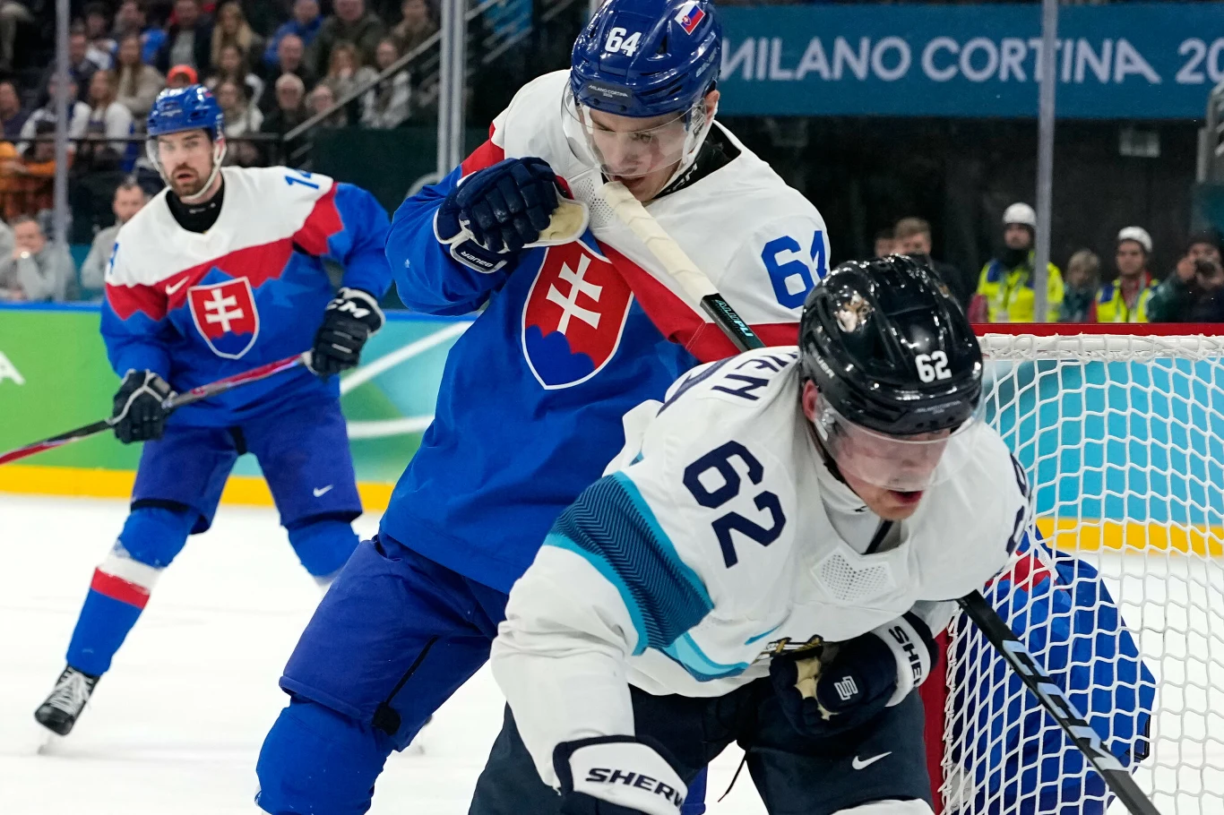 Dwóch hokeistów walczy o krążek podczas meczu, zawodnik w niebiesko-czerwonym stroju z numerem 64 reprezentuje Słowację, natomiast zawodnik w białym stroju z niebieskimi elementami ma numer 62, w tle widoczny jest tłum kibiców.