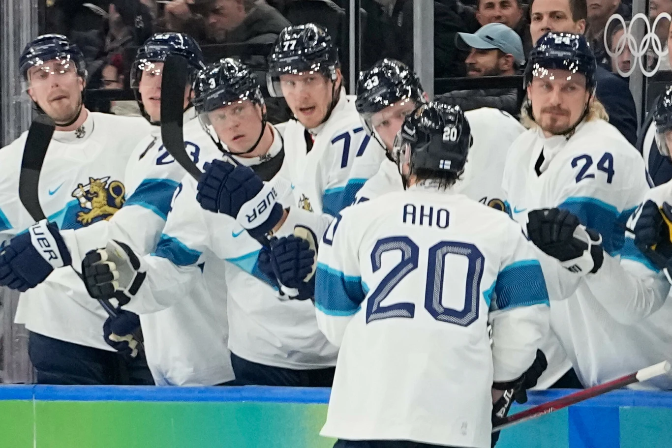 Zawodnicy drużyny hokejowej ubrani w białe stroje z niebieskimi akcentami, świętujący przy ławce po udanej akcji jednego z graczy o nazwisku Aho i numerze 20. W tle widzowie oraz logo olimpijskie.