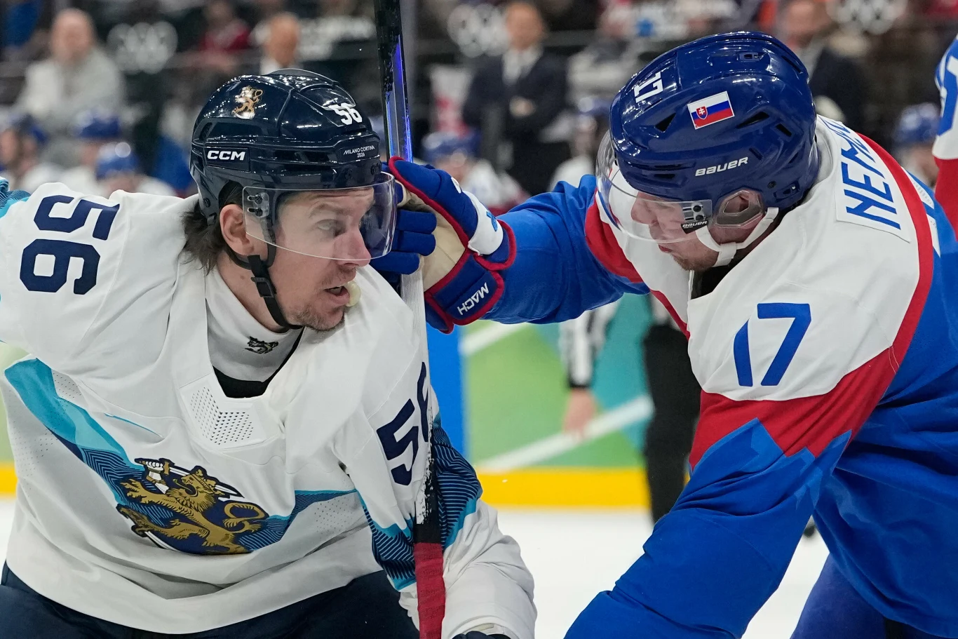 Dwóch hokeistów w trakcie zaciętej walki o krążek, jeden w białym stroju z numerem 56 i z herbem lwa, drugi w niebiesko-białej koszulce z numerem 17 i słowacką flagą na kasku. Zawodnik w niebiesko-białym stroju delikatnie dotyka twarzy przeciwnika ręką...