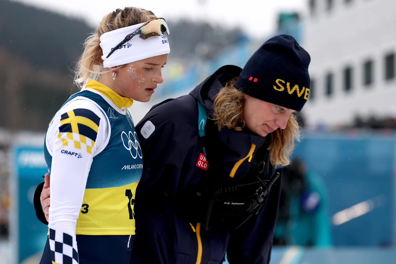 Frida Karlsson Dwie osoby w sportowych strojach, jedna z nich ubrana w strój narciarski i opaskę na głowie, druga w ciemnej kurtce z napisem SWEB na czapce, stoją razem, okazując wsparcie lub otuchę. W tle rozmyty budynek oraz elementy toru narciarskiego.