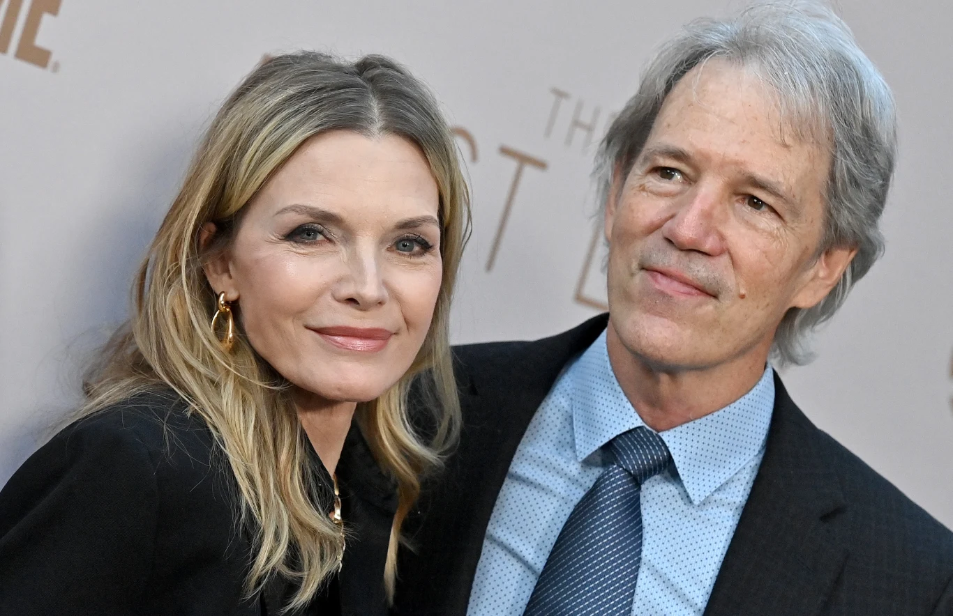 Dwie dorosłe osoby stojące obok siebie ubrane elegancko, kobieta w czarnym stroju z jasnymi włosami, mężczyzna w garniturze i niebieskiej koszuli, oboje uśmiechnięci na tle jasnego, neutralnego tła.