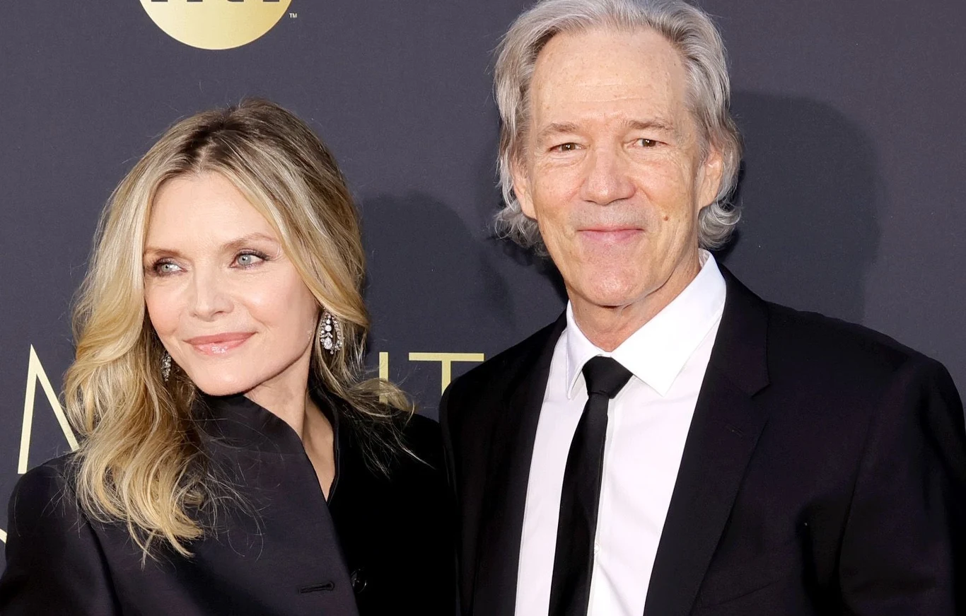 Dwoje elegancko ubranych osób pozuje razem na tle ciemnej ścianki z napisem, kobieta o jasnych włosach uśmiecha się, a mężczyzna z siwymi włosami stoi obok niej w garniturze.