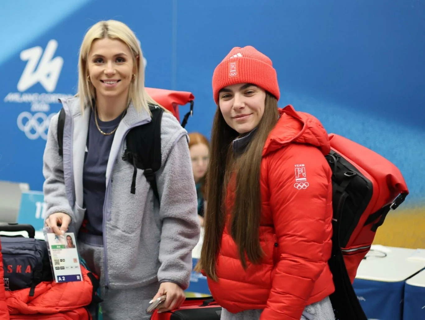 Linda Weiszewski (po prawej) i Klaudia Adamek Dwie młode kobiety w sportowych strojach zimowych, jedna w jasnej bluzie i spodniach, druga w czerwonej kurtce i czapce z emblematem olimpijskim oraz napisem Polska, stoją z plecakami na tle niebieskiej, olimpijskiej scenerii.
