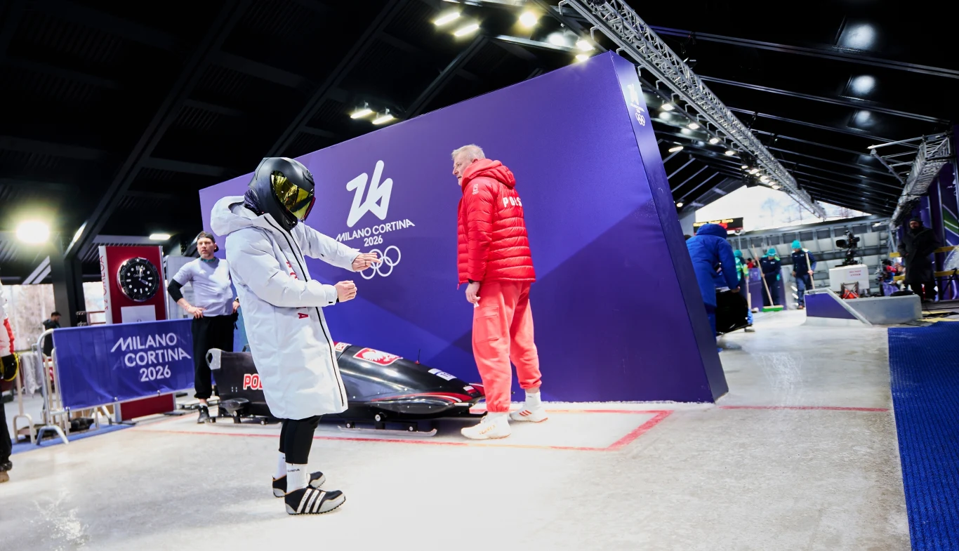 Zawodnik w stroju ochronnym przygotowuje się do startu przy bobsleju na tle baneru Igrzysk Olimpijskich Mediolan Cortina 2026, obok instruktor w czerwonej kurtce. W tle widać sprzęt sportowy i elementy zaplecza technicznego.