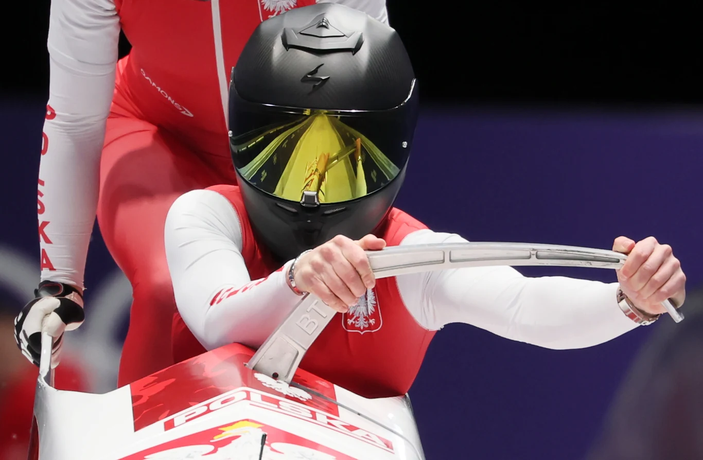 Zawodnik ubrany w czerwono-biały kombinezon z polskimi emblematami, siedzący w bobsleju i trzymający kierownicę, z kaskiem chroniącym głowę, przygotowujący się do sportowego zjazdu.