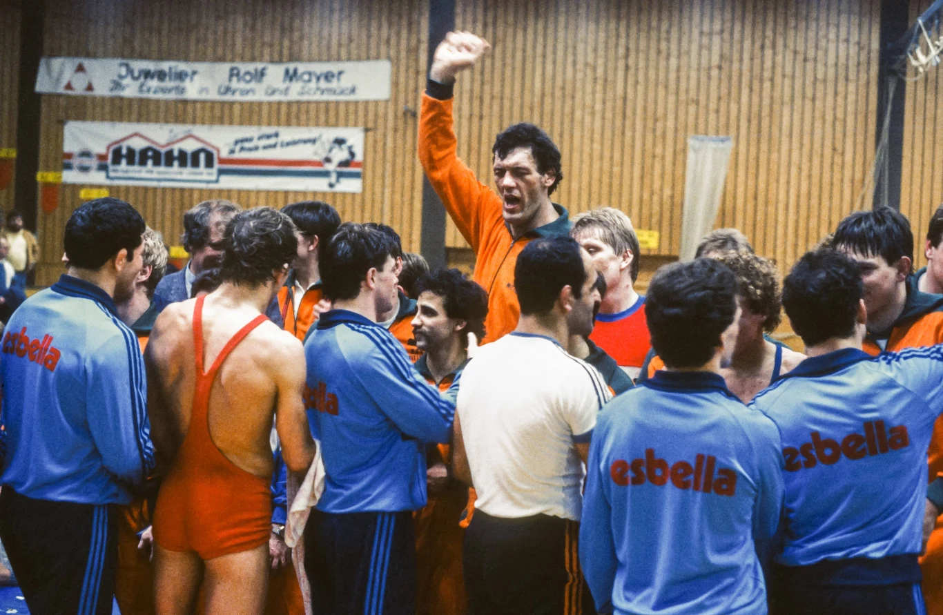 Grupa sportowców zgromadzonych wokół jednej osoby unoszącej rękę w geście zwycięstwa lub mobilizacji. Zawodnicy ubrani są w sportowe stroje w odcieniach niebieskim, czerwonym i pomarańczowym, otoczeni halą sportową z drewnianymi ścianami i reklamami kl...