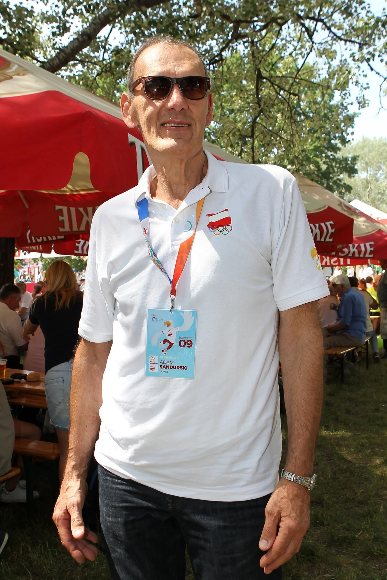 Mężczyzna w okularach przeciwsłonecznych i białej koszulce polo z identyfikatorem zawieszonym na szyi stoi na tle namiotu z logotypem, w otoczeniu innych ludzi siedzących przy stołach plenerowych.