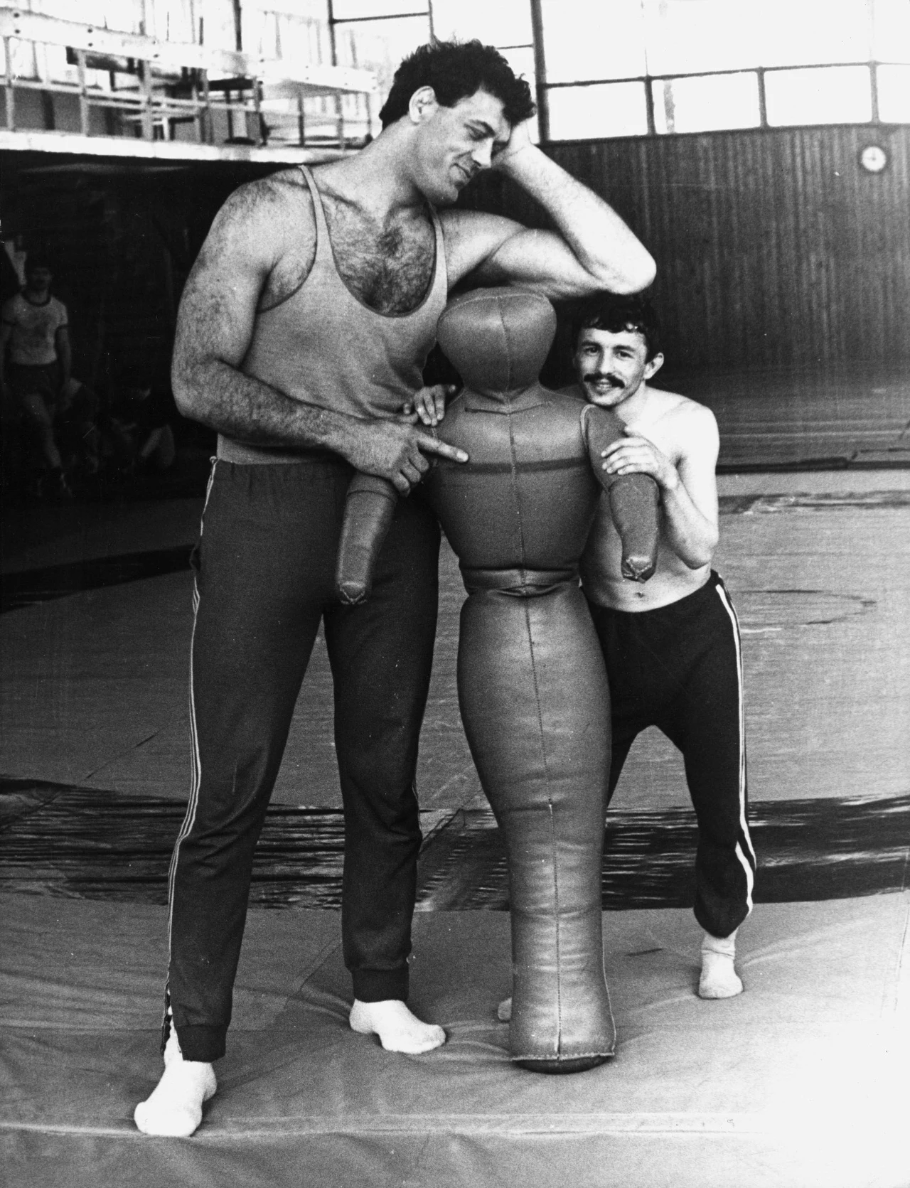 Dwóch mężczyzn o wyraźnie różnym wzroście w sportowych strojach trenuje z manekinem w sali gimnastycznej. Wyższy mężczyzna opiera się swobodnie o głowę manekina, podczas gdy niższy obejmuje go od boku.