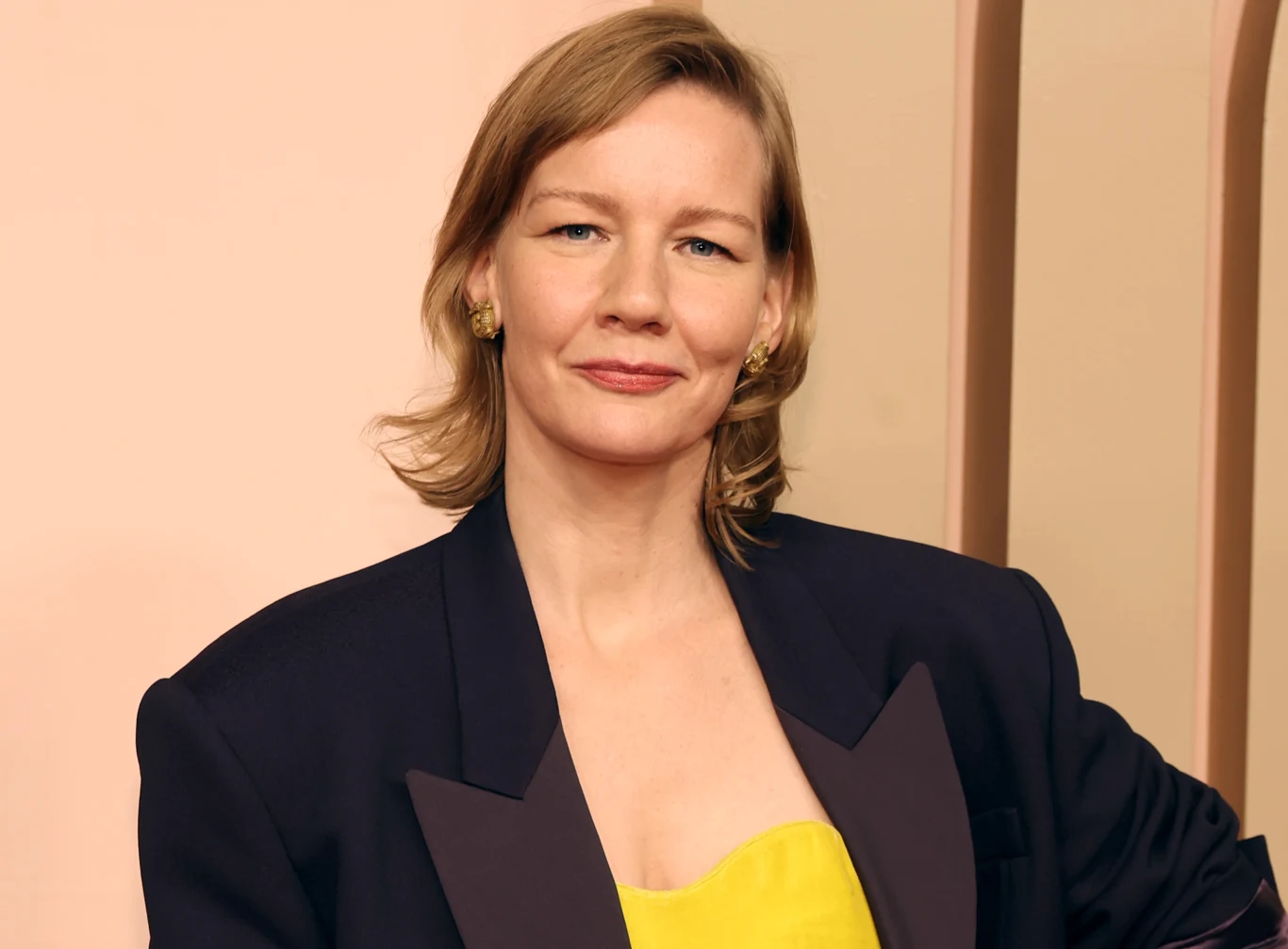 Sandra Huller Kobieta o blond włosach, ubrana elegancko w ciemną marynarkę i żółtą sukienkę, stoi na jasnym tle z delikatnym uśmiechem i pewną siebie postawą.