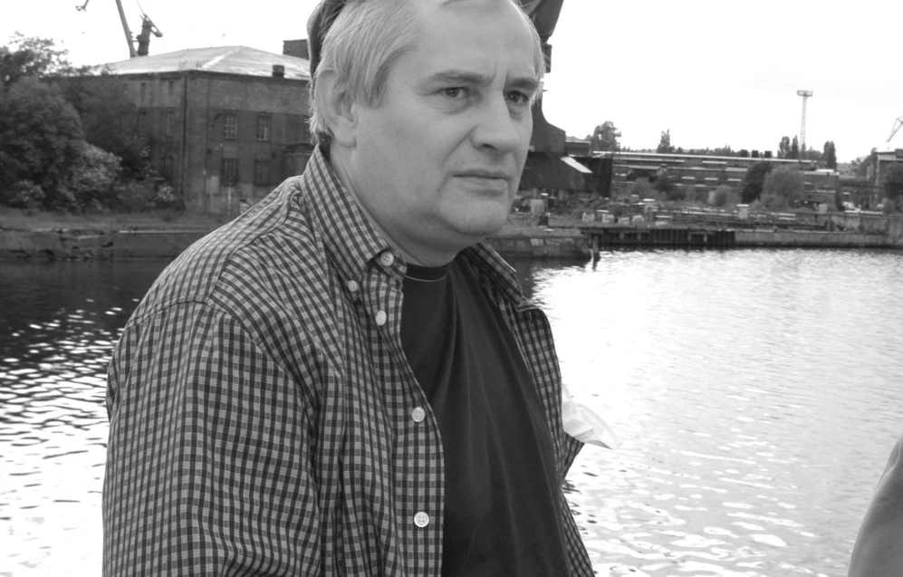 Mirosław Krawczyk Mężczyzna w średnim wieku stoi nad wodą, ubrany w koszulę w kratę i ciemny podkoszulek. W tle widoczny jest industrialny krajobraz z budynkami, żurawiami i linią brzegową.