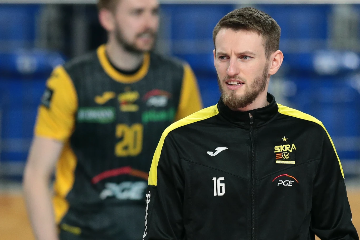 Kacper Piechocki przez blisko dekadę grał w Bełchatowie Zawodnik drużyny PGE Skra Bełchatów w czarno-żółtej kurtce sportowej stoi na hali sportowej, w tle widoczny drugi siatkarz w stroju meczowym.