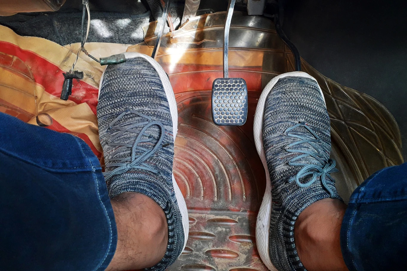 Sportowe buty na nogach osoby siedzącej na miejscu kierowcy. Na podłodze znajdują się pedały sprzęgła i hamulca oraz wycieraczka samochodowa o wzorze w czerwono-brązowe łuki.