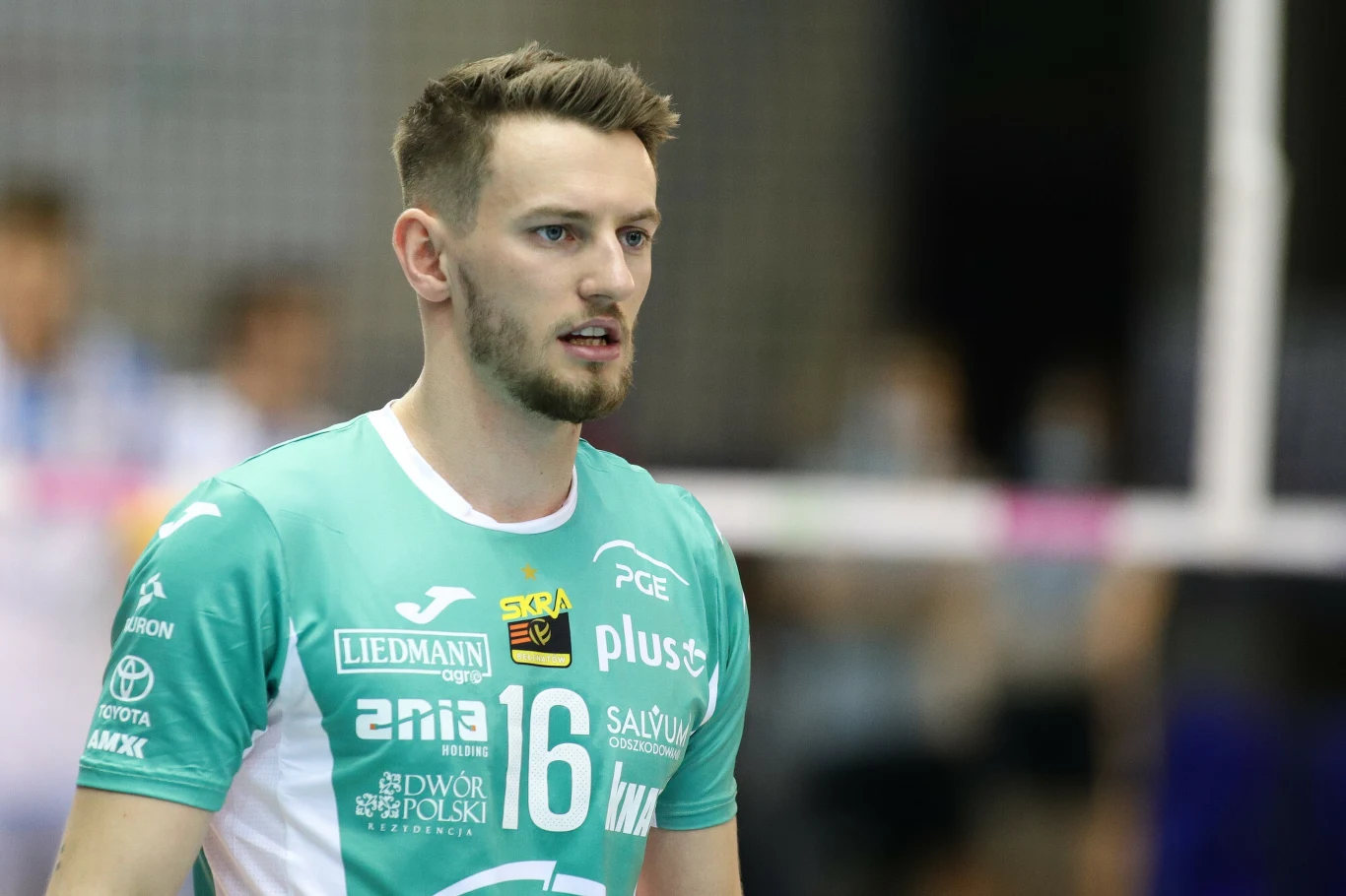 Kacper Piechocki wrócił do wydarzeń sprzed lat. Opowiedział o rozstaniu ze Skrąe Młody mężczyzna w sportowej koszulce siatkarskiej z numerem 16, stoi na hali sportowej i skupia wzrok przed siebie, w tle widoczne rozmazane postacie oraz elementy siatki.