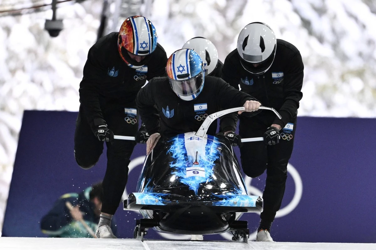 Skandal podczas zawodów bobslejowych Trzech sportowców w kombinezonach i kaskach popycha bobslej, każdy z nich ma na kasku flagę Izraela, tło stanowi zimowy pejzaż i elementy toru bobslejowego.