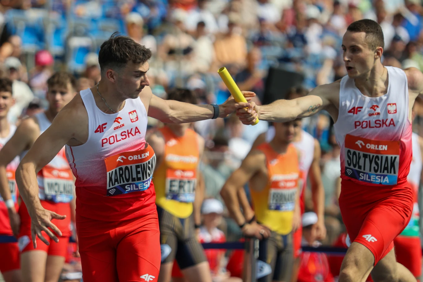 Daniel Sołtysiak i Marcin Karolewski Dwaj polscy lekkoatleci w biało-czerwonych strojach przekazują sobie pałeczkę podczas biegu sztafetowego na stadionie, w tle widoczni inni zawodnicy oraz trybuny z kibicami.