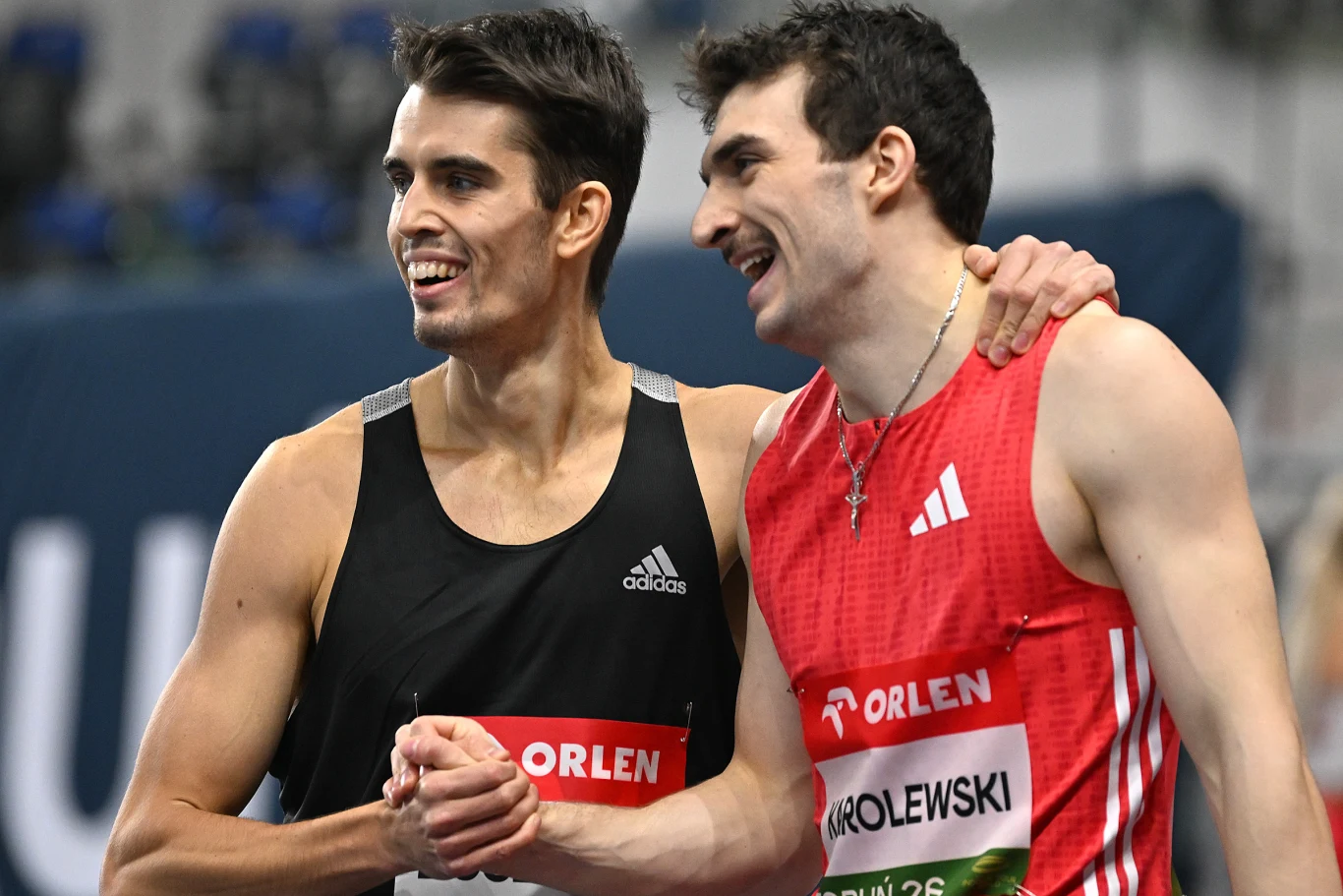 Kajetan Duszyński (z lewej) i Marcin Karolewski Dwóch sportowców w strojach z numerami startowymi obejmuje się i uśmiecha na tle hali sportowej, okazując radość i koleżeństwo po zakończonych zawodach.