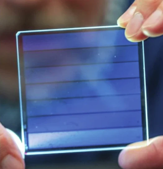 Przezroczysty panel słoneczny trzymany pomiędzy palcami dwóch dłoni, delikatnie odbijający światło, wyraźnie widoczne poziome linie wskazują na technologię cienkowarstwową.