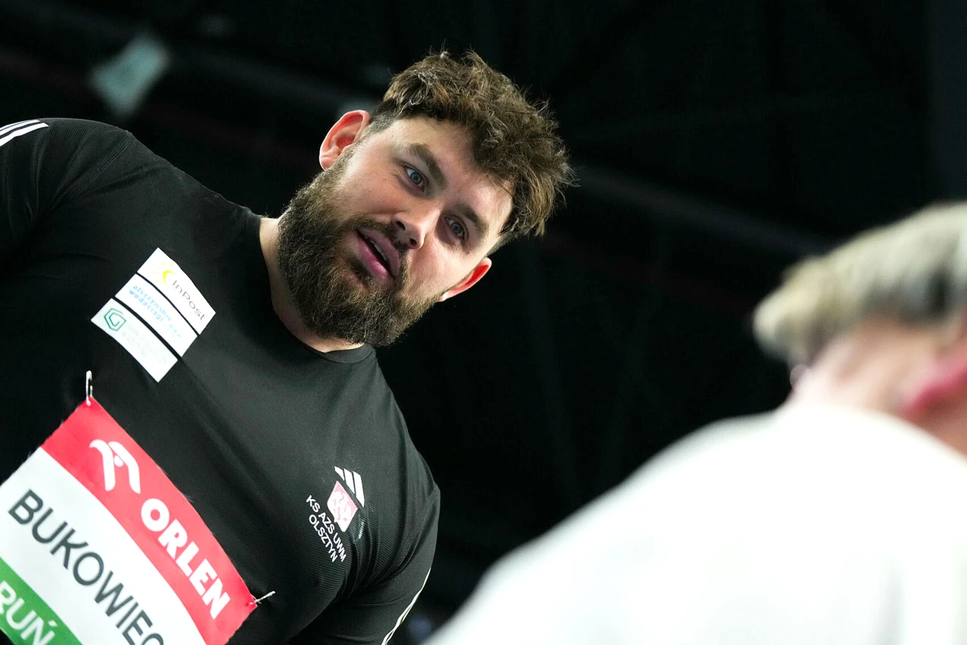 Konrad Bukowiecki Mężczyzna z brodą w czarnej koszulce sportowej z identyfikatorem z nazwiskiem, zbliżenie na twarz. W tle niewyraźna sylwetka innej osoby, scena odbywa się najprawdopodobniej na hali sportowej.