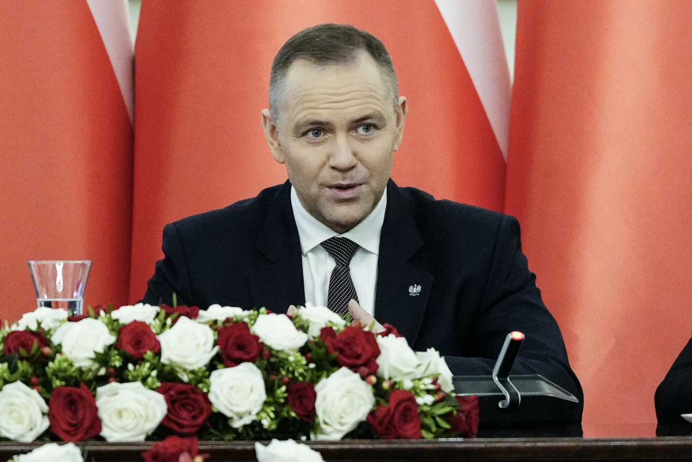 “Godna emerytura” prezydenta w Sejmie. Projekt budzi obawy Mężczyzna w garniturze siedzi przy stole ozdobionym bukietem biało-czerwonych róż, za nim widać tło złożone z polskich flag.