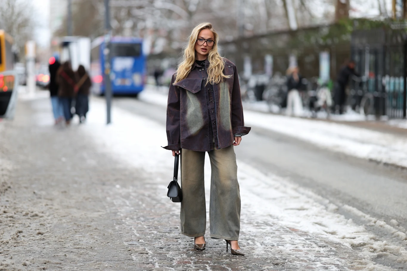 Kobieta w okularach o długich blond włosach stoi na ośnieżonym chodniku ubrana w szerokie spodnie, ciemną kurtkę i szpilki, trzymając w ręku czarną torebkę; w tle widać autobusy i zimową ulicę.