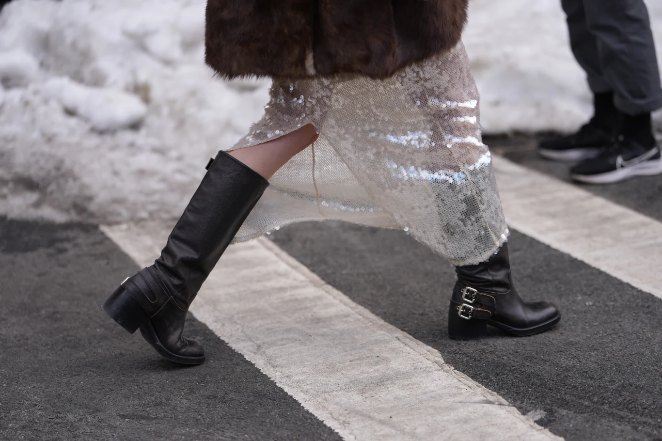 Czarne wysokie skórzane kozaki na grubym obcasie, cekinowa spódnica oraz futrzana kurtka widoczne w ruchu na przejściu dla pieszych; w tle śnieg.