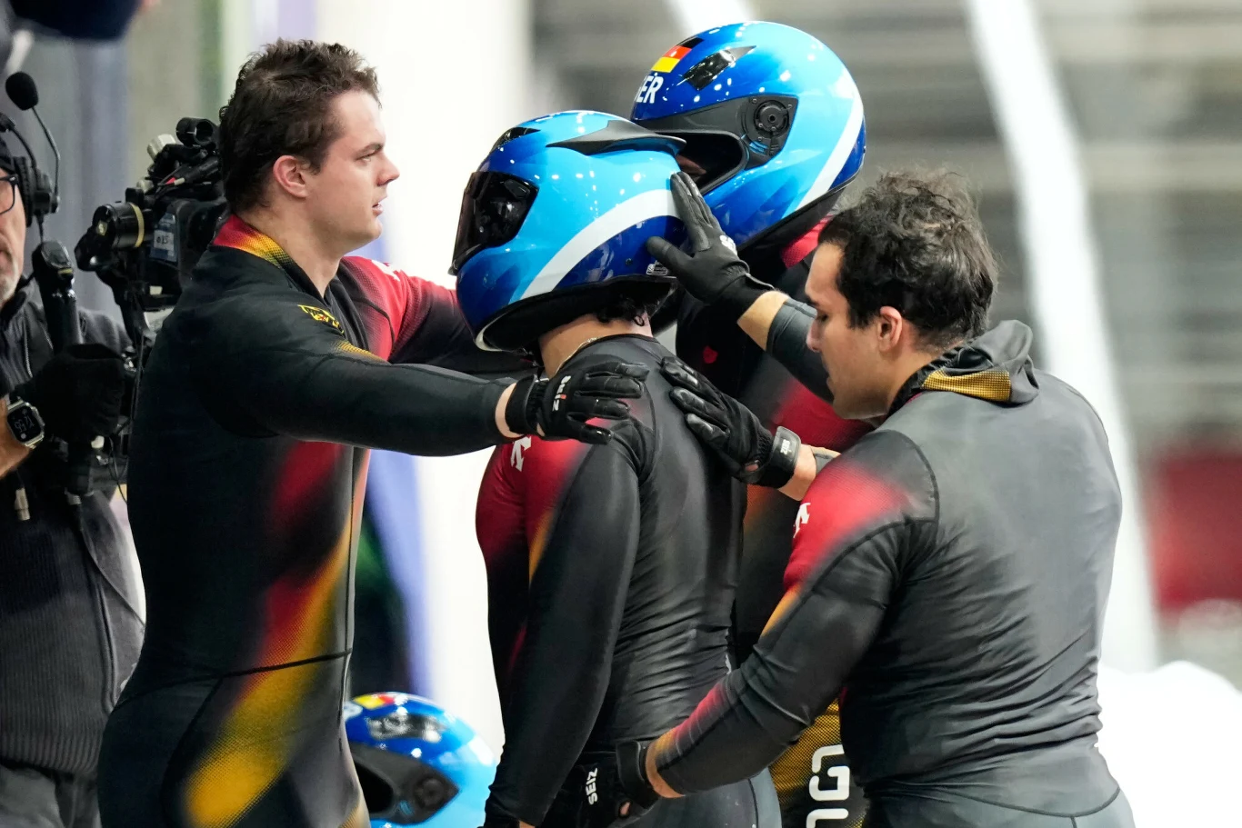 Ekipa Adama Ammoura podczas rywalizacji na igrzyskach Zawodnicy drużyny bobslejowej w kombinezonach i niebieskich kaskach przygotowują się do startu lub kończą przejazd, jeden z nich jest otoczony przez pozostałych, co sugeruje chwilę wsparcia lub współpracy sportowej.