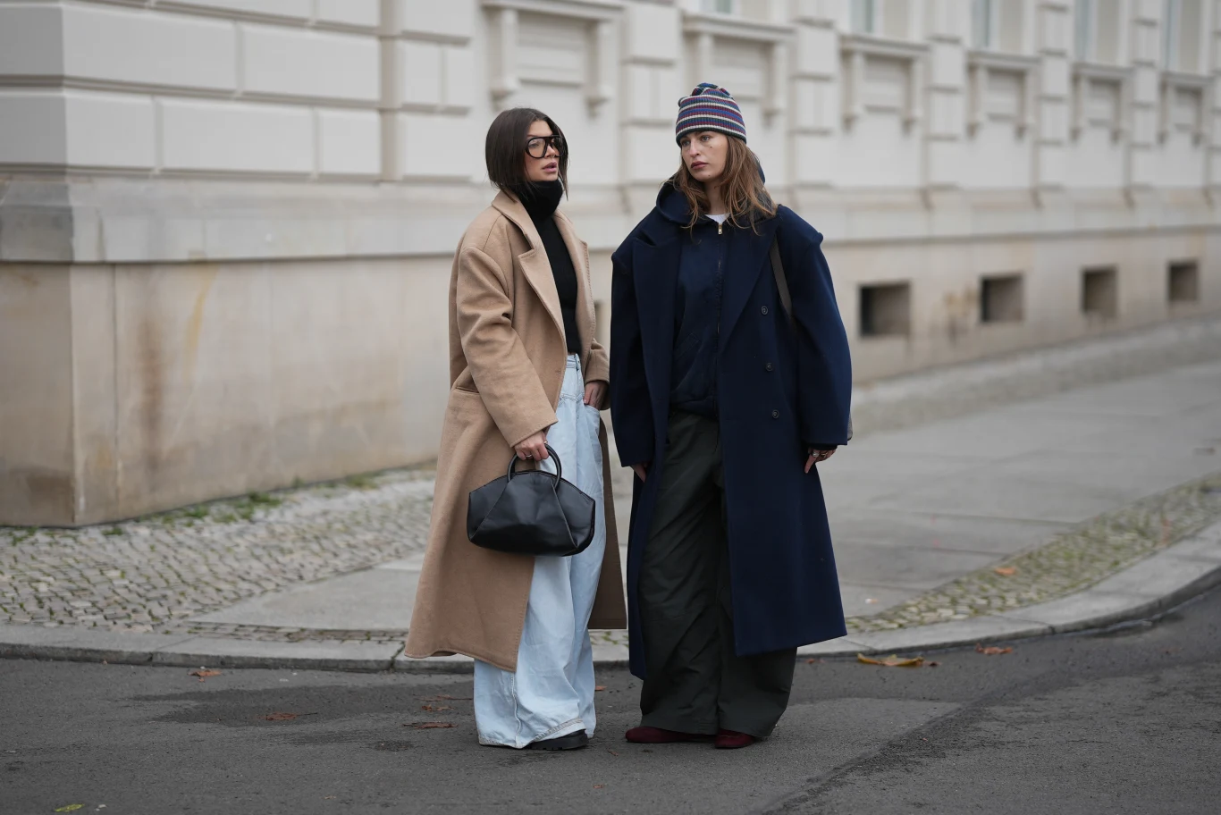 Dwie kobiety ubrane w modne płaszcze stoją na chodniku przy ulicy miejskiej, jedna z nich trzyma czarną torebkę, obie wyróżniają się stylowymi zimowymi stylizacjami, jedna z nich nosi czapkę w paski.