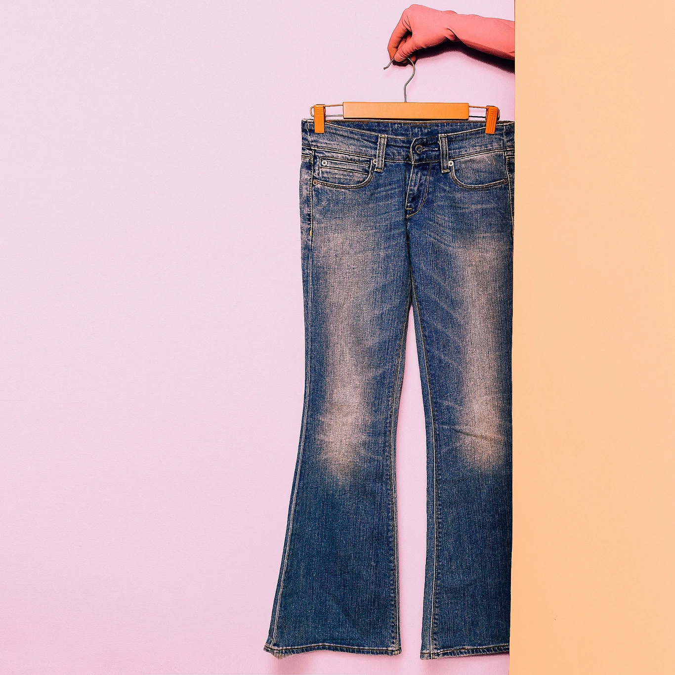 Bootcut to model jeansów z lekko poszerzaną nogawką Dżinsowe spodnie na wieszaku trzymane przez rękę wyłaniającą się zza żółtej ścianki, na tle jasnofioletowej ściany.