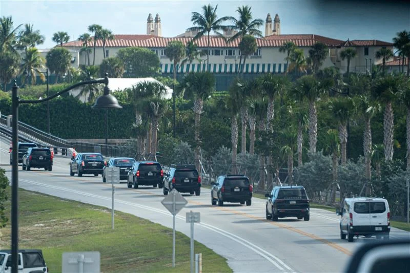 Kolumna czarnych samochodów typu SUV jedzie drogą otoczoną palmami, w tle widać dużą rezydencję z czerwonym dachem oraz palmami, po lewej stronie lampy uliczne, po prawej biały van.