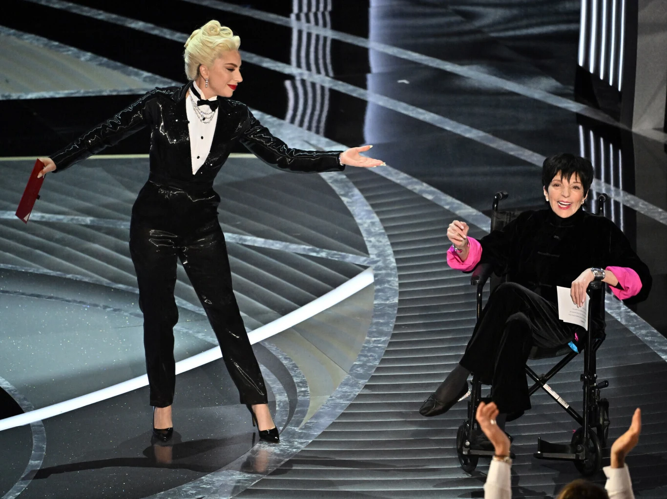 Lady Gaga i Liza Minnelli podczas rozdania Oscarów w 2022 roku Elegancko ubrana kobieta w garniturze stojąca na scenie wykonuje gest w kierunku drugiej kobiety, siedzącej na wózku inwalidzkim i trzymającej kopertę, obie uśmiechnięte, wokół widoczne elementy scenografii w stylu gali.