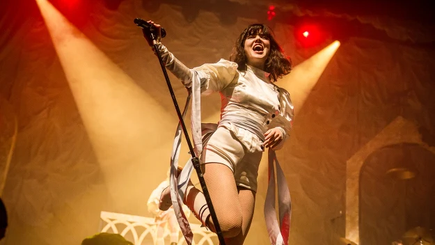 Wokalistka występująca na oświetlonej scenie podczas koncertu, trzymająca mikrofon i ubrana w biały kostium sceniczny. W tle widoczny perkusista oraz scenografia z łukami architektonicznymi, całość podkreślona ciepłym światłem reflektorów.