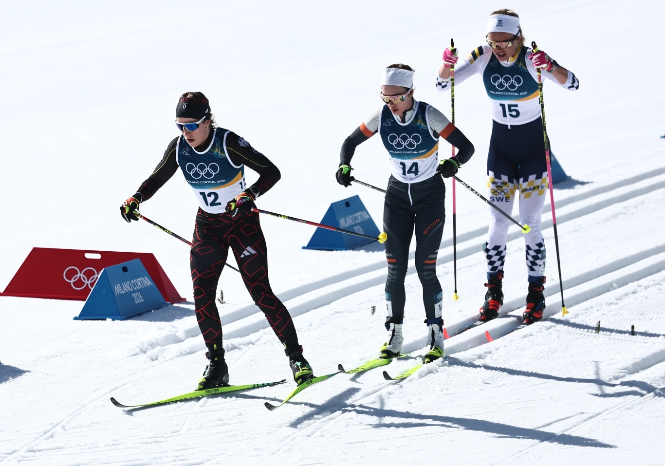 Trzy kobiety w sportowych strojach rywalizują w biegu narciarskim podczas Zimowych Igrzysk Olimpijskich, poruszając się po wyznaczonej trasie na tle śnieżnego krajobrazu, widoczne tablice olimpijskie i numery startowe.