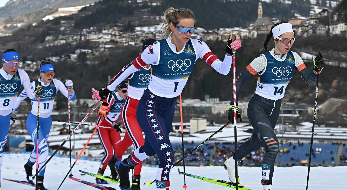 Zawodniczki rywalizujące w biegu narciarskim podczas zimowych igrzysk olimpijskich, ubrane w stroje sportowe z numerami startowymi, poruszają się po śnieżnej trasie na tle górskiego krajobrazu i zabudowań.