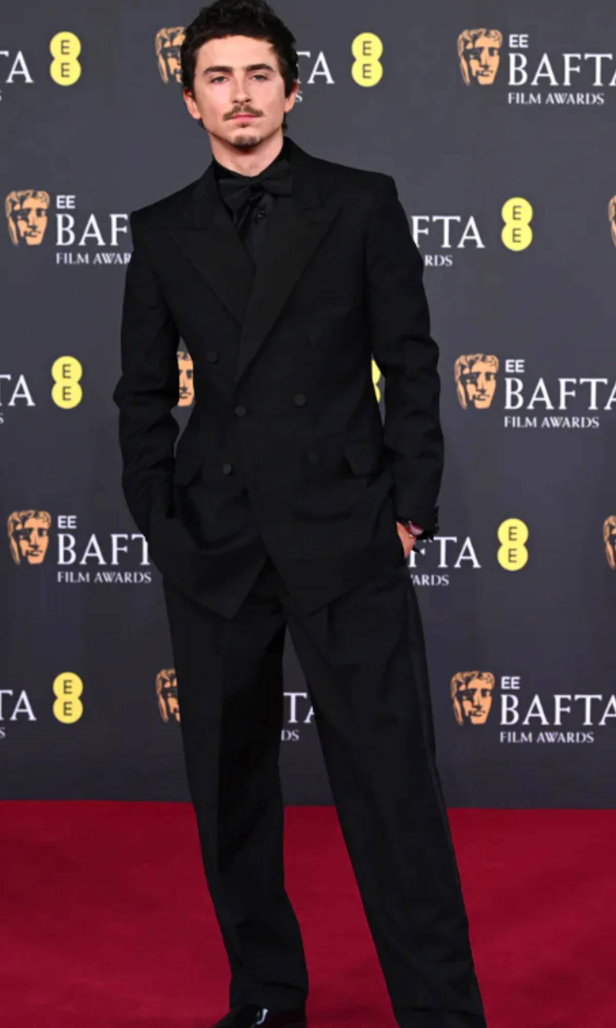 Elegancko ubrany młody mężczyzna w czarnym garniturze stojący na czerwonym dywanie na tle ściany z logotypami BAFTA.