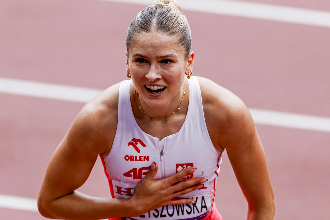Pia Skrzyszowska Sportsmenka w stroju lekkoatletycznym z logo Orlen i godłem Polski, stoi na bieżni, uśmiecha się i trzyma dłoń na klatce piersiowej. Jej wyraz twarzy wskazuje na silne emocje, prawdopodobnie po zakończeniu wyścigu.