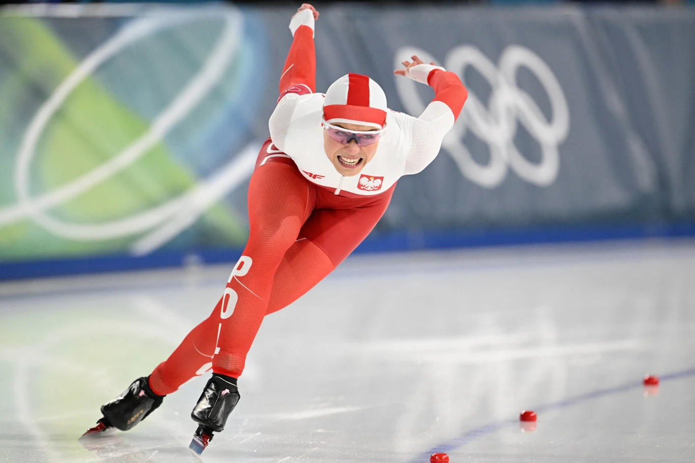 Łyżwiarz szybki w biało-czerwonym stroju z godłem Polski na piersi dynamicznie pokonuje zakręt na lodowisku olimpijskim, z wyraźną determinacją na twarzy.