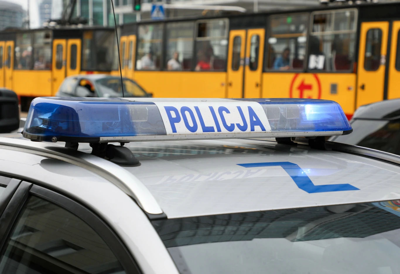 Tragedia w tramwaju (zdj. ilustr.) Policyjny radiowóz z włączonymi niebieskimi światłami ostrzegawczymi na tle przejeżdżającego żółtego tramwaju w przestrzeni miejskiej.