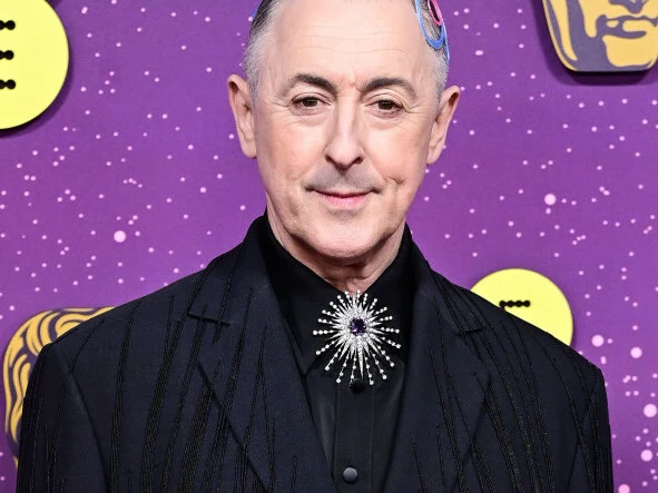 Alan Cumming podczas 79. ceremonii rozdania nagród BAFTA Mężczyzna w eleganckim, czarnym garniturze z ozdobą w kształcie gwiazdy na kołnierzu, pozujący na tle fioletowej ściany ozdobionej żółtymi i białymi elementami graficznymi.