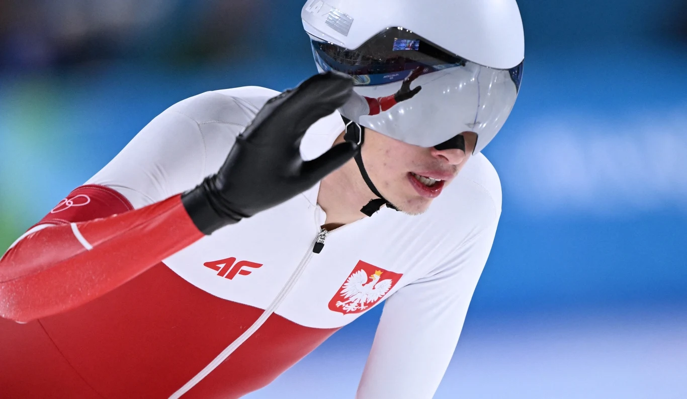 Zawodnik w kombinezonie sportowym reprezentującym Polskę podczas wyścigu łyżwiarskiego, ujęcie dynamiczne, widoczne lustrzane gogle, gest ręką w trakcie jazdy.