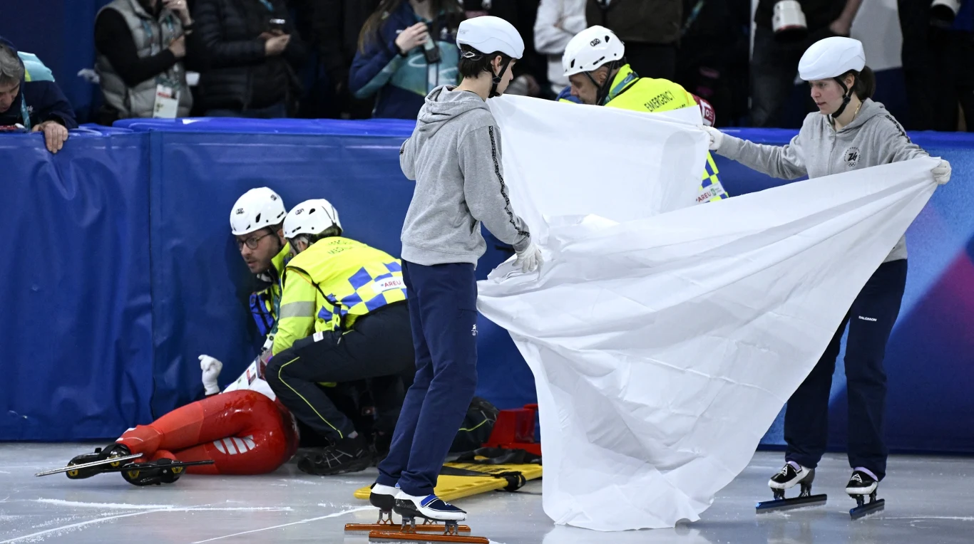 Zawodnik leży na lodowisku w pozycji sugerującej kontuzję, obok niego klęczy ratownik medyczny, a dwóch pomocników trzyma białe prześcieradło, zasłaniając widok dla publiczności; wszyscy noszą kaski ochronne