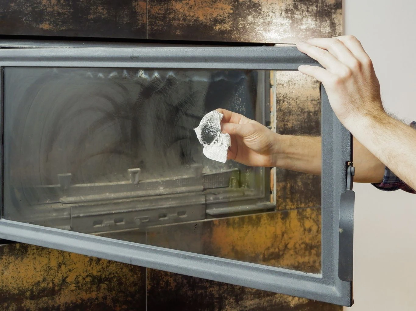 Ręka osoby czyści zabrudzoną szybę kominka papierowym ręcznikiem, wnętrze kominka lekko zakurzone, a ściany w tle mają metaliczną strukturę.
