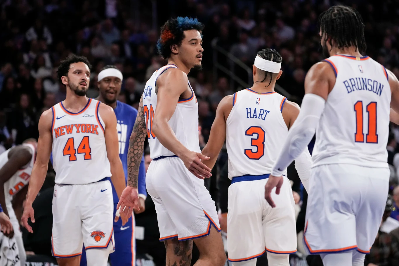 Koszykarze drużyny New York Knicks ubrani w białe stroje sportowe podczas meczu na parkiecie, wśród nich kilku zawodników odwróconych plecami z widocznymi nazwiskami na koszulkach, tłum w tle.