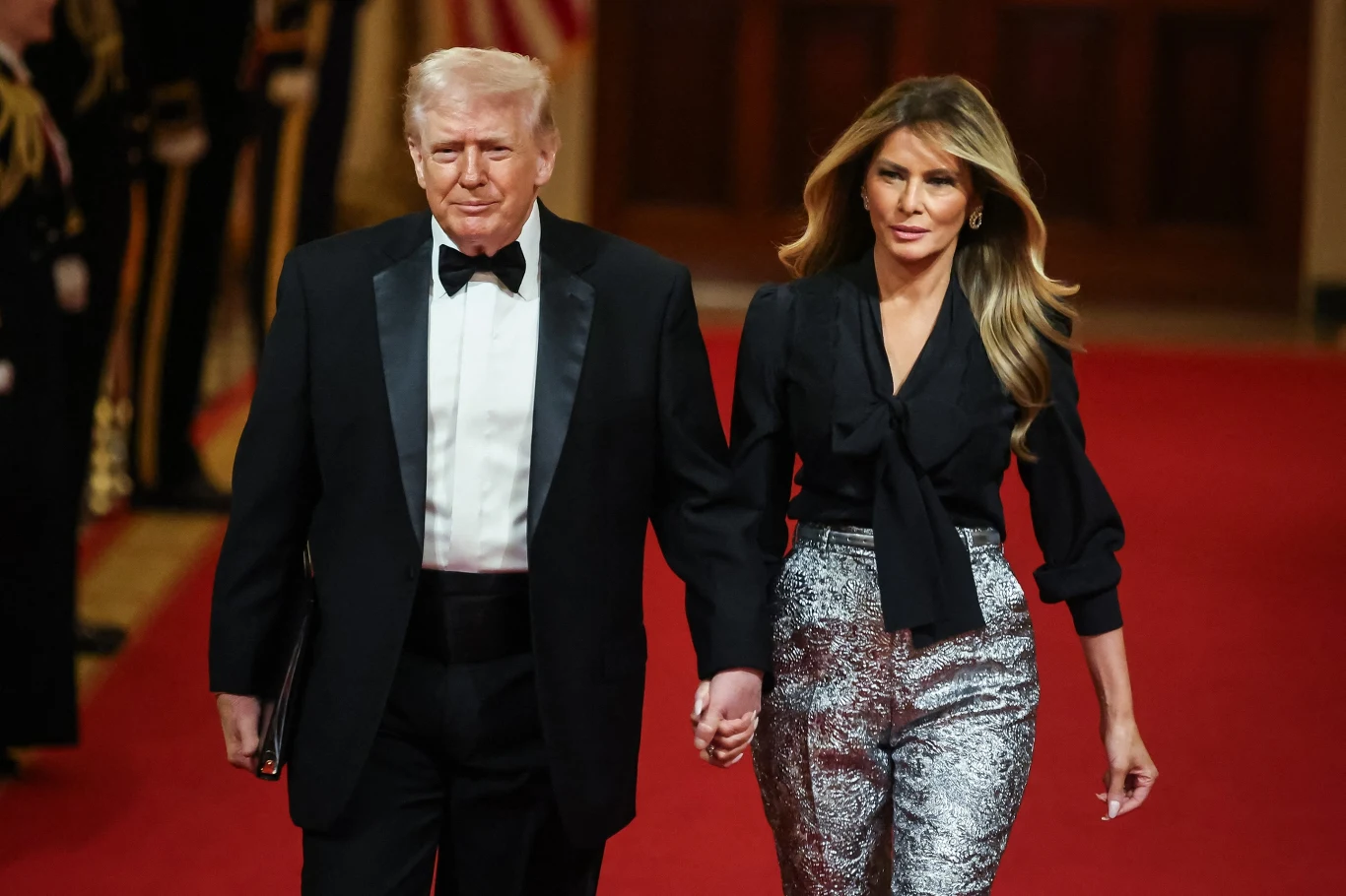 Melania Trump błyszczała u boku męża podczas kolacji Mężczyzna w smokingu i kobieta w eleganckim stroju idą razem po czerwonym dywanie, trzymając się za ręce, w tle widać osoby w mundurach i eleganckie wnętrze.