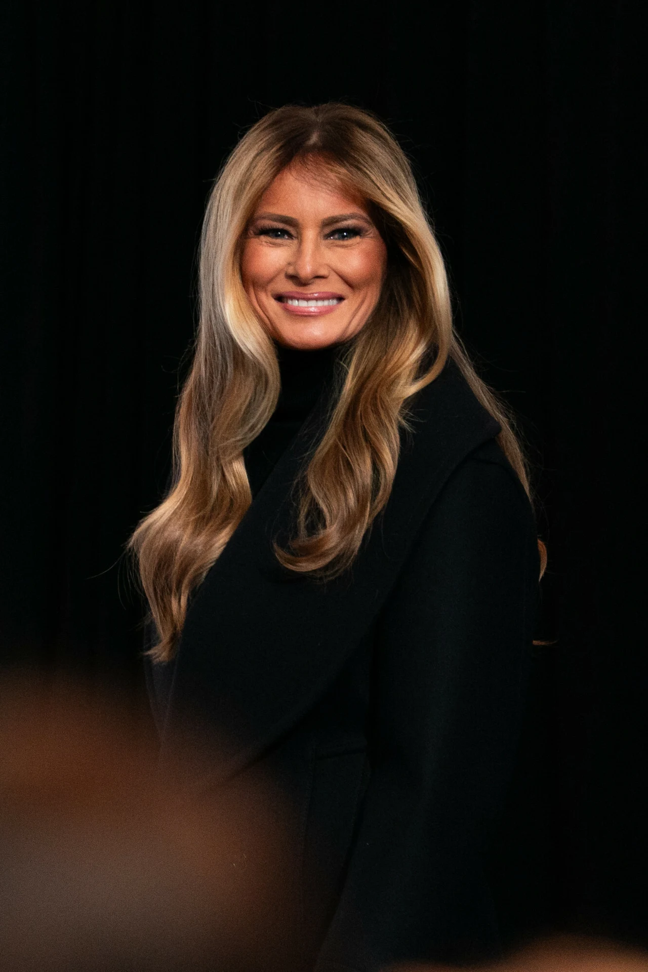 Włosy Melanii są częścią jej wizerunku, który inspiruje inne kobiety Uśmiechnięta kobieta z długimi, jasnobrązowymi włosami, ubrana w elegancką, czarną garderobę, stoi na ciemnym tle.