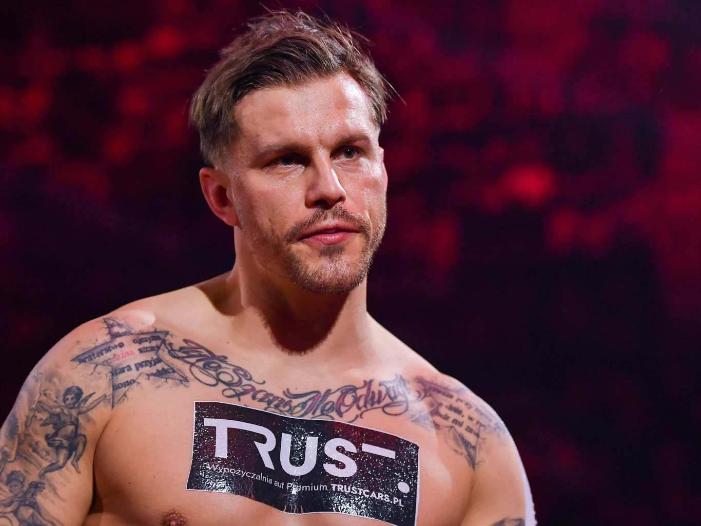Alan Kwieciński Mężczyzna o umięśnionej sylwetce z licznymi tatuażami na torsie stoi na tle czerwonego światła, na piersi ma naklejkę z logo firmy Trust.
