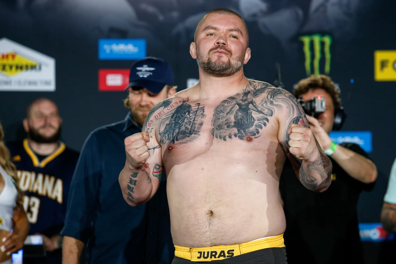 Mężczyzna o muskularnej sylwetce stoi w pozie zaciśniętych pięści, eksponując tatuaże na klatce piersiowej oraz brzuchu, z wyrazem pewności siebie na twarzy. W tle kilka osób patrzy w jego stronę, część z nich patrzy w obiektyw, a inni są zajęci swoimi...